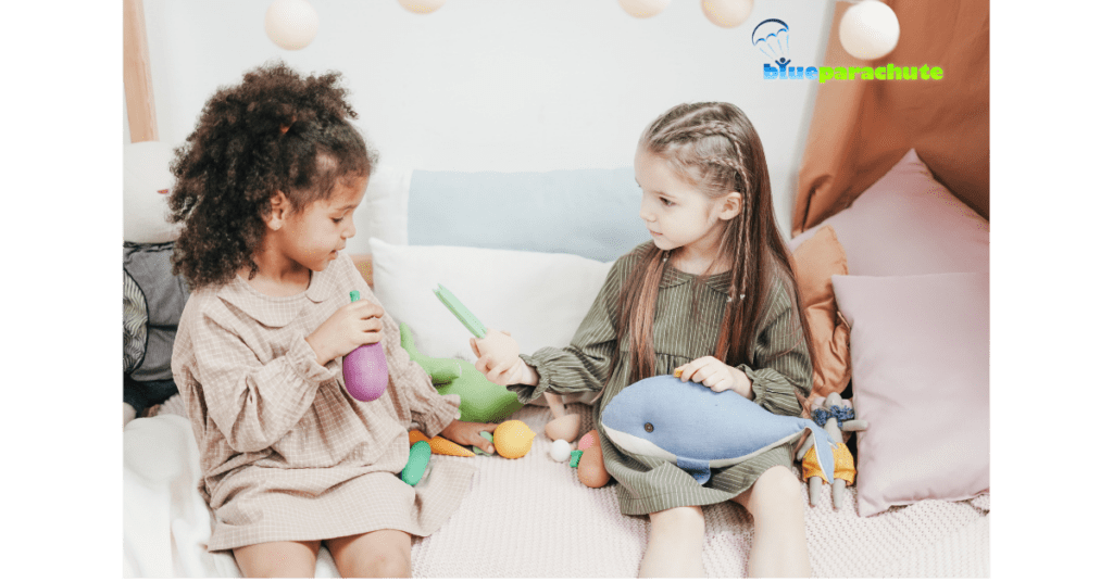 Two young girls sitting on a couch, and one of the young girls is holding a play vegetable and extending her arm towards the other girl. This implies she is learning to share.