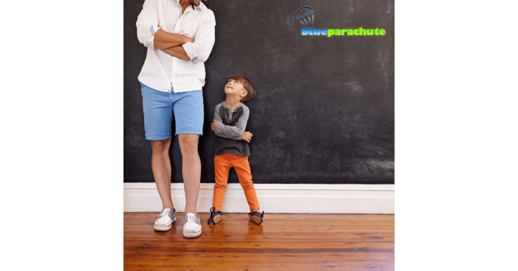 A father and a son, standing against a wall, with the son looking up at the dad. They both have their arms crossed, indicating that the son is imitating the dad. This implies teaching your child to learn by imitation.