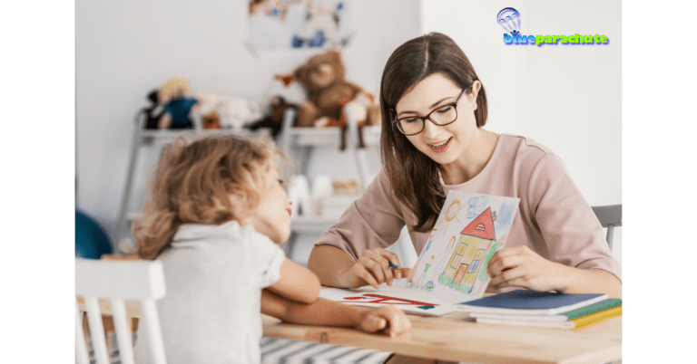 A female teacher who is about 25 years old and wearing glasses is showing a young child, who is sitting across the table from her, a hand-drawn picture of a house with people next to the house and the sun in the sky. This exemplifies how she is using effective autism teaching strategies.